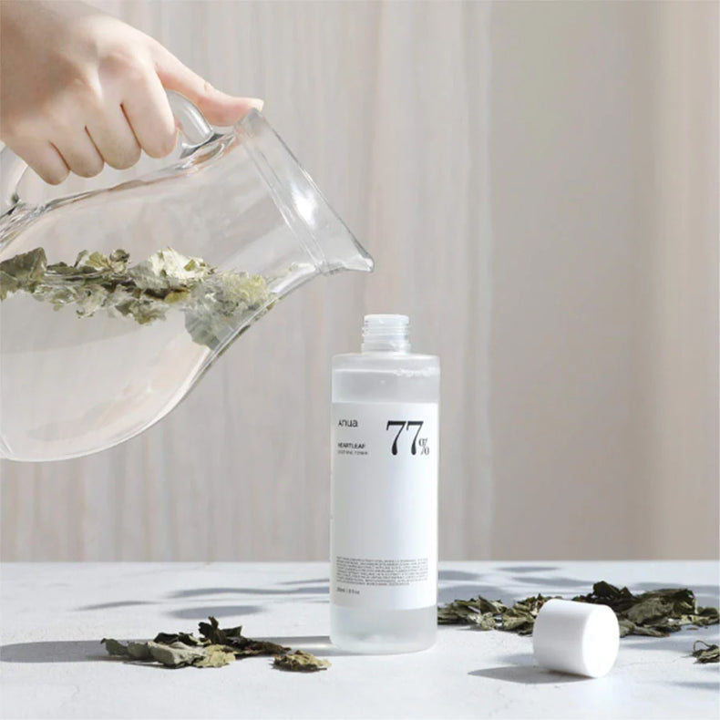 Anua Heartleaf 77% Soothing Toner bottle with the cap off, as water with leaves is poured from a glass pitcher nearby.