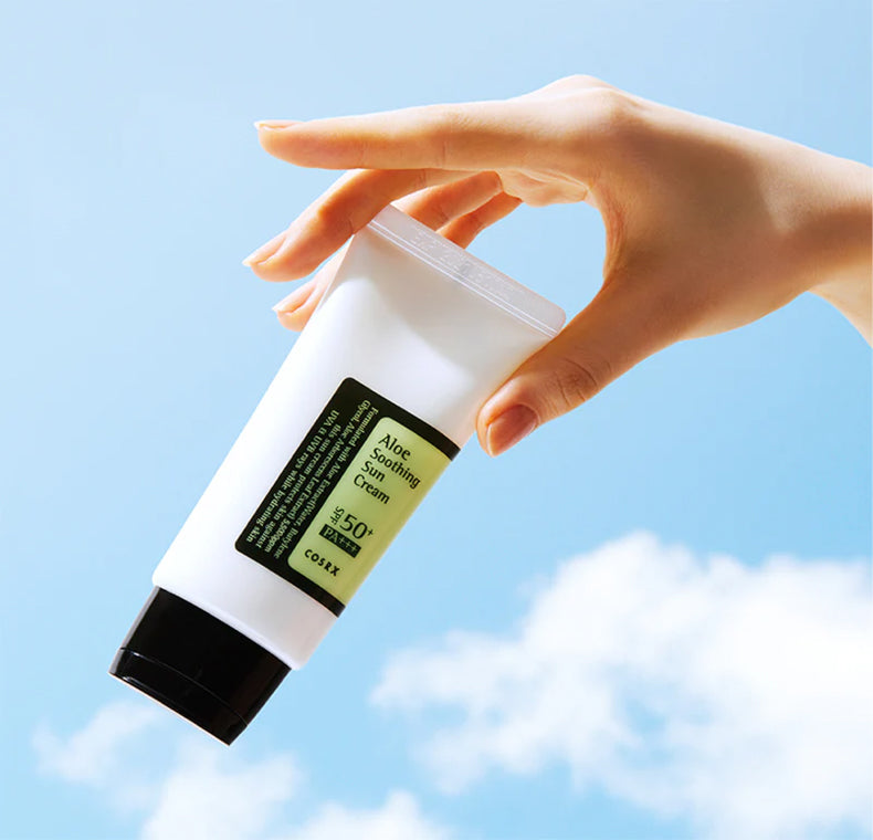 Hand holding a tube of COSRX Aloe Soothing Sun Cream SPF 50+ PA+++ against a bright blue sky with clouds.