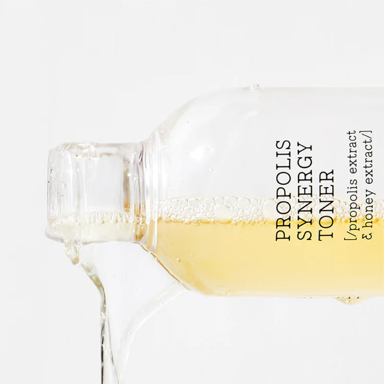 Close-up of COSRX Propolis Synergy Toner bottle tilted, showing the golden liquid pouring out with bubbles visible inside