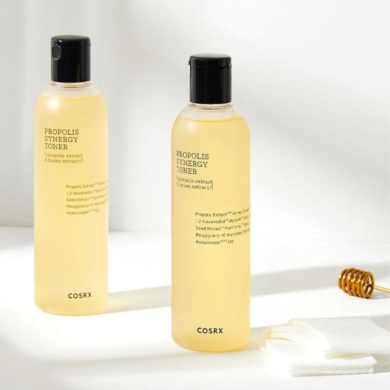 Two bottles of COSRX Propolis Synergy Toner standing on a light surface, accompanied by cotton pads and a honey dipper.