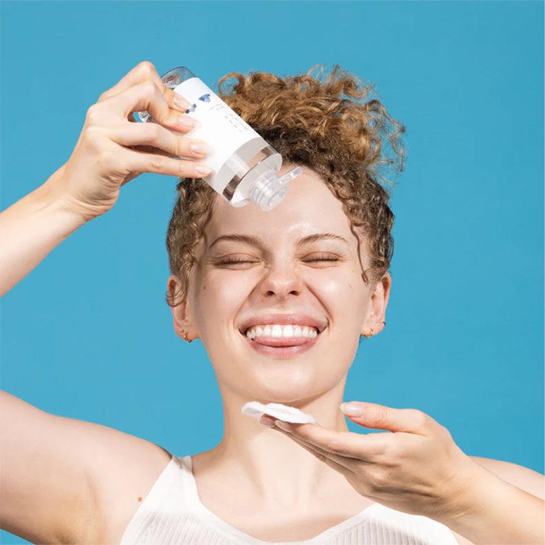 Smiling woman applying Round Lab 1025 Dokdo Toner with a cotton pad against a blue background.
