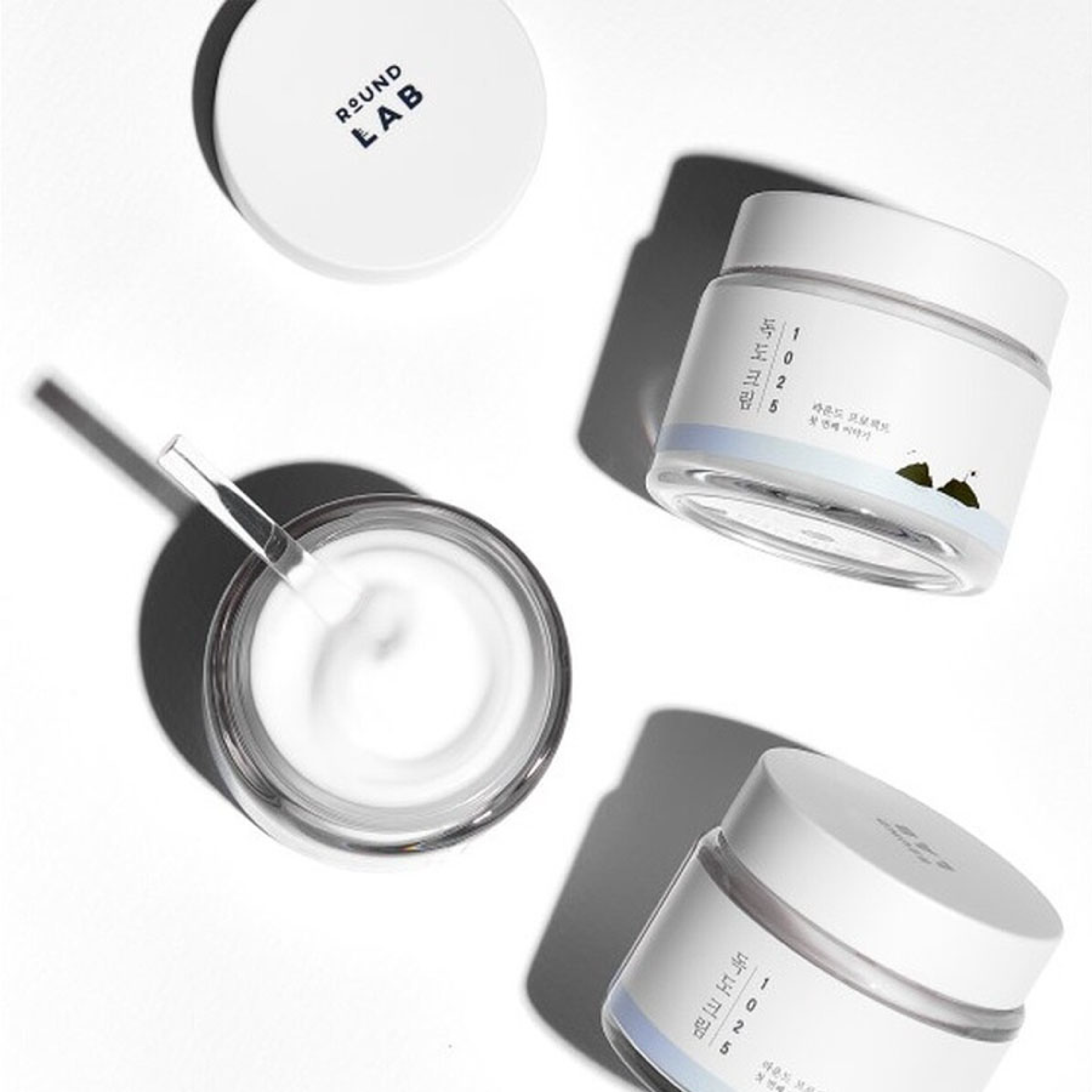 Three jars of Round Lab 1025 Dokdo Cream, one open with a spatula, arranged on a white surface.