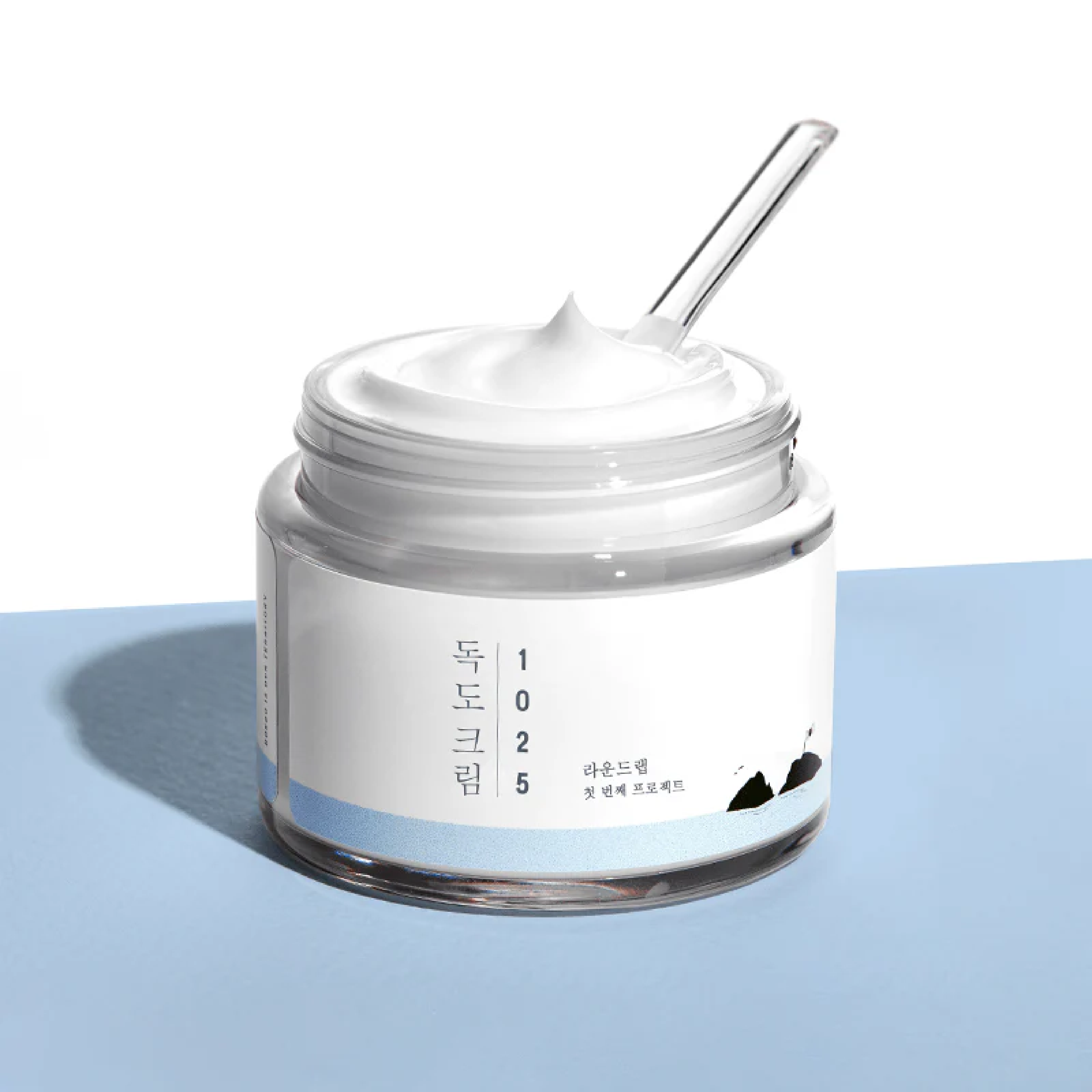 Open jar of Round Lab 1025 Dokdo Cream with a spatula, on a blue surface.